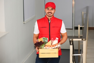 Photo of Male courier holding wooden crate with products indoors. Food delivery service