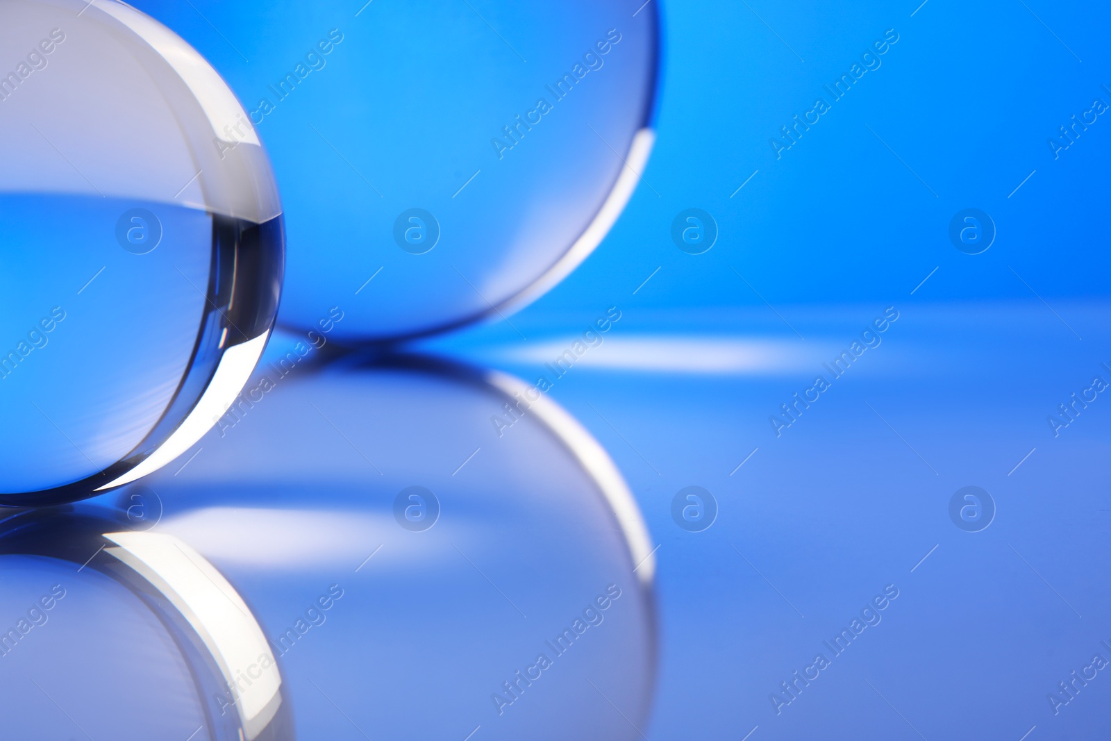 Photo of Transparent glass balls on mirror surface against blue background, closeup