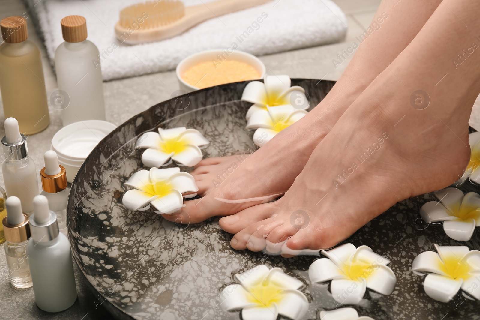 Photo of Woman soaking her feet in bowl with water and flowers on floor, closeup. Spa treatment