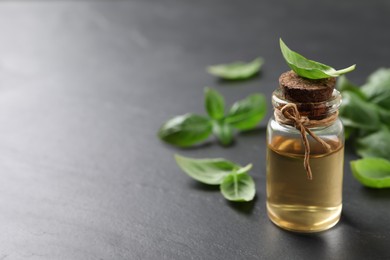 Photo of Bottle of essential basil oil and fresh leaves on dark grey table, space for text