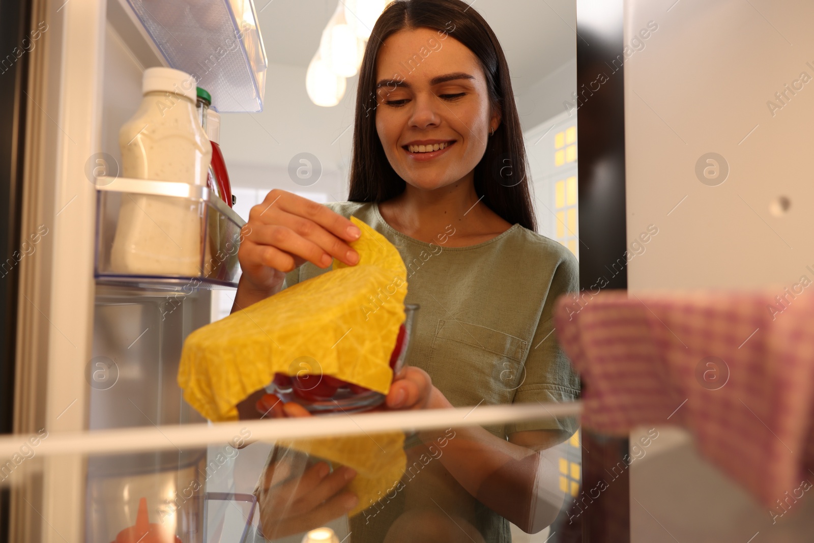 Photo of Happy woman taking away beeswax food wrap, view from refrigerator