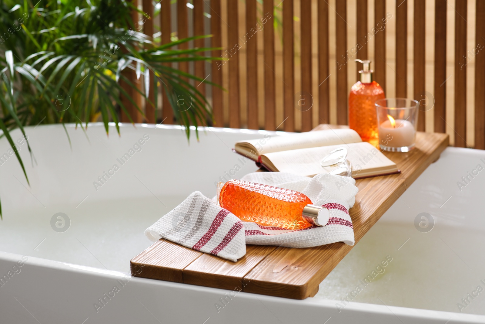 Photo of Wooden bath tray with open book, candle and body care products on tub indoors. Relaxing atmosphere