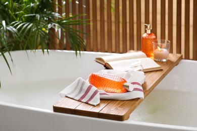 Photo of Wooden bath tray with open book, candle and body care products on tub indoors. Relaxing atmosphere