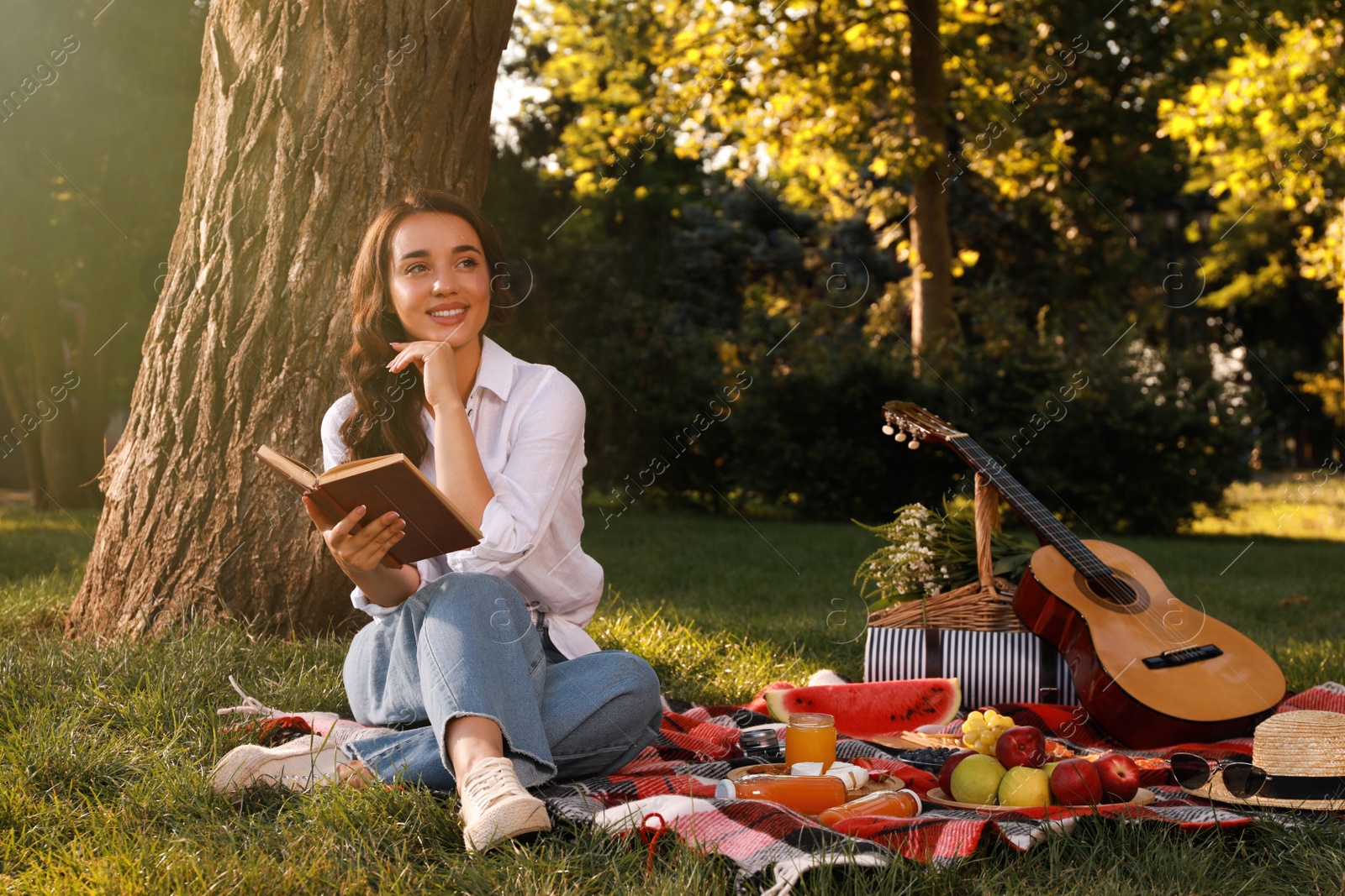 Photo of Happy young woman reading book on plaid in park. Summer picnic