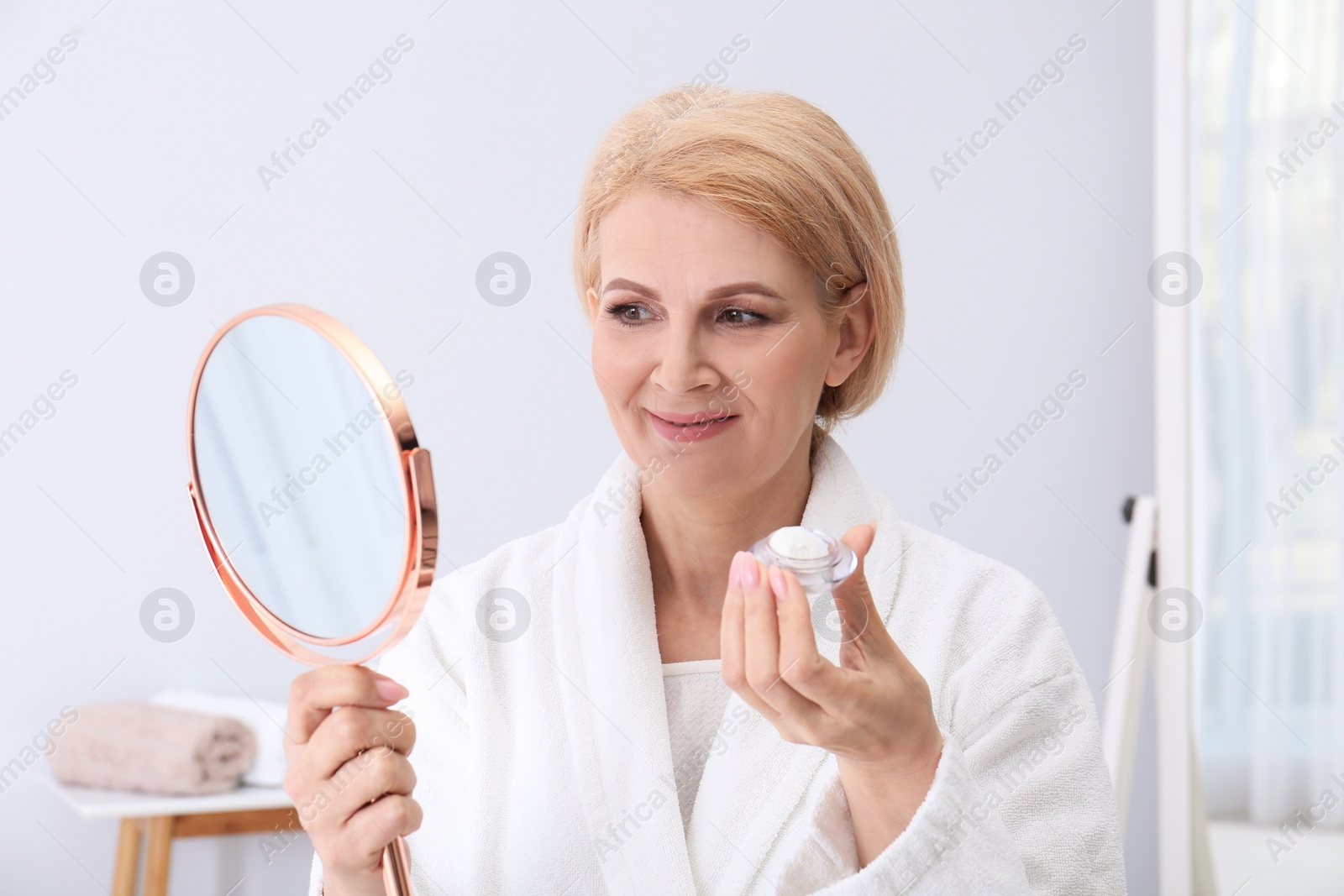 Photo of Mature woman with jar of face cream and mirror at home