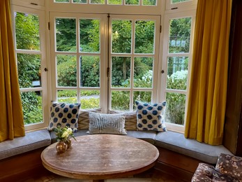 Wooden table near bay window seat with cushions in cozy room. Interior design