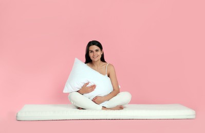 Photo of Young woman sitting on soft mattress and holding pillow against pink background