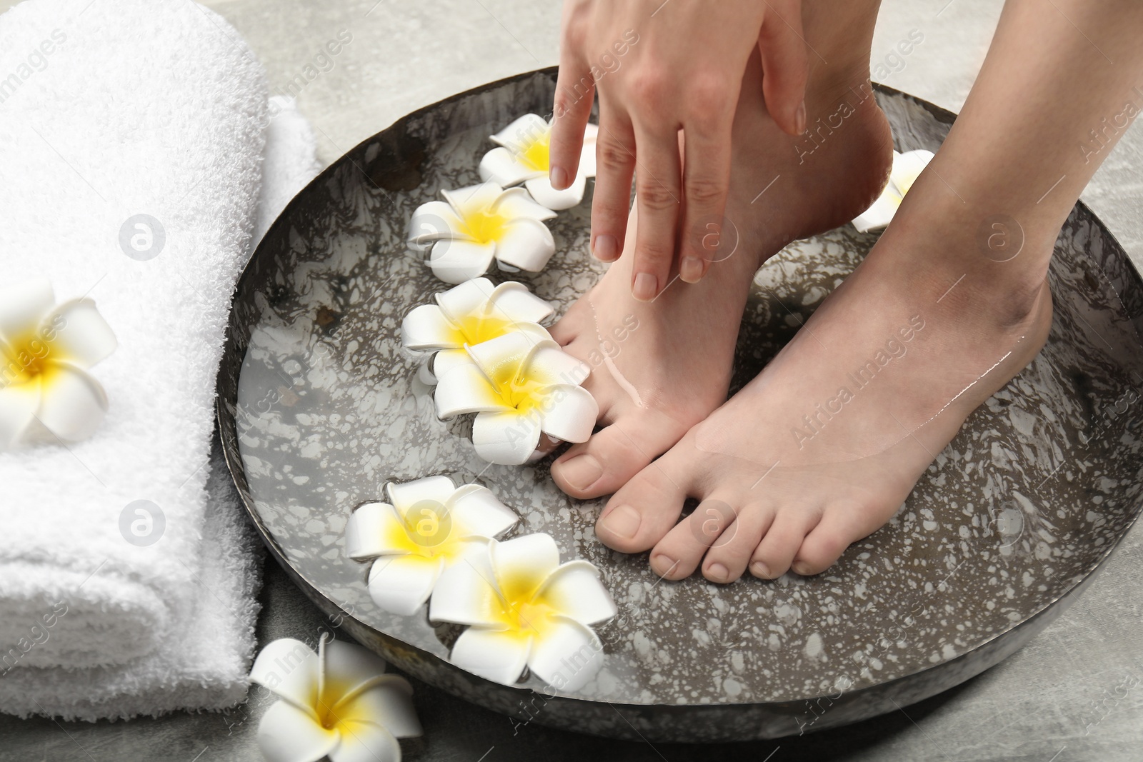 Photo of Woman soaking her feet in bowl with water and flowers on floor, closeup. Spa treatment