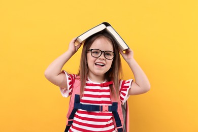 Photo of Cute little girl in glasses with open book and backpack against orange background