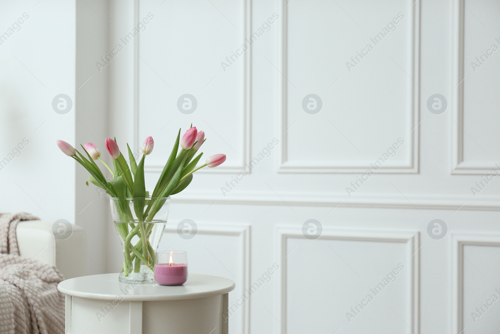 Photo of Beautiful tulips and burning candle on white table indoors, space for text. Interior design