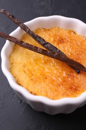 Photo of Delicious creme brulee in bowl and vanilla pods on dark gray textured table, closeup
