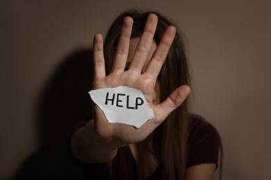 Abused young woman with sign HELP near beige wall, focus on hand. Domestic violence concept