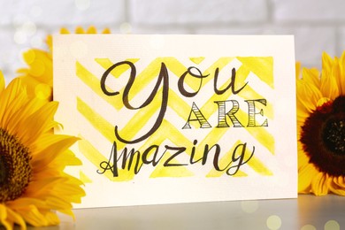 Card with life-affirming phrase You Are Amazing and sunflowers on light table