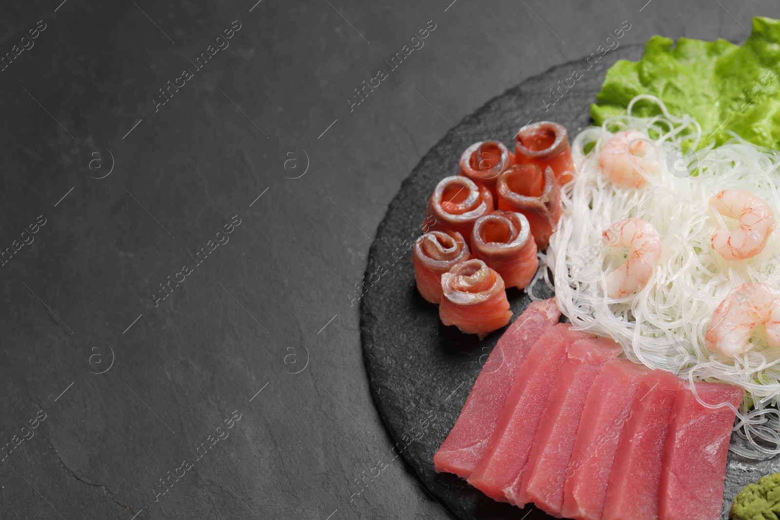 Photo of Sashimi set (raw tuna, salmon slices and shrimps) served with funchosa, lettuce and vasabi on dark table, above view. Space for text
