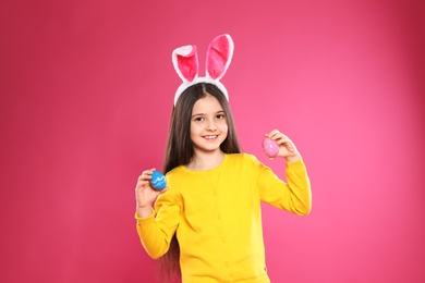 Little girl in bunny ears headband holding Easter eggs on color background