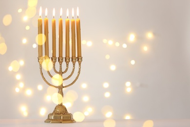 Photo of Golden menorah with burning candles against light grey background and blurred festive lights, space for text