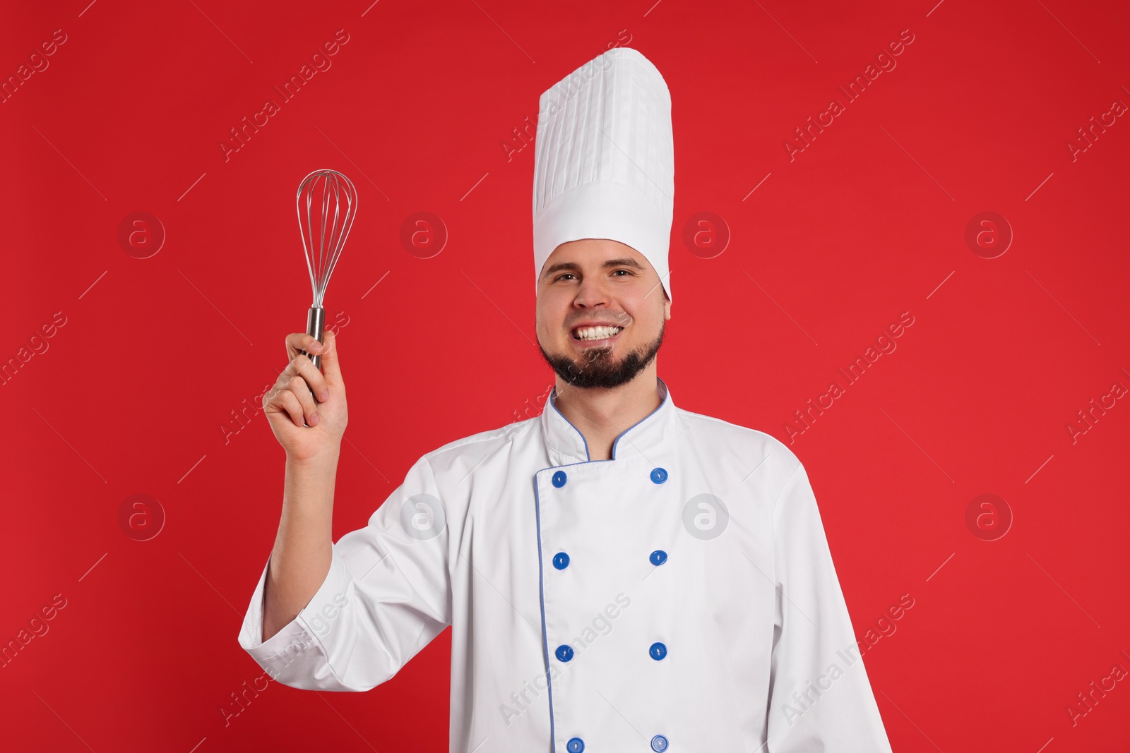 Photo of Happy professional confectioner in uniform holding whisk on red background