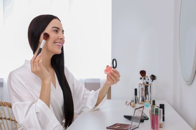 Photo of Beautiful young woman with blusher and brush at dressing table indoors
