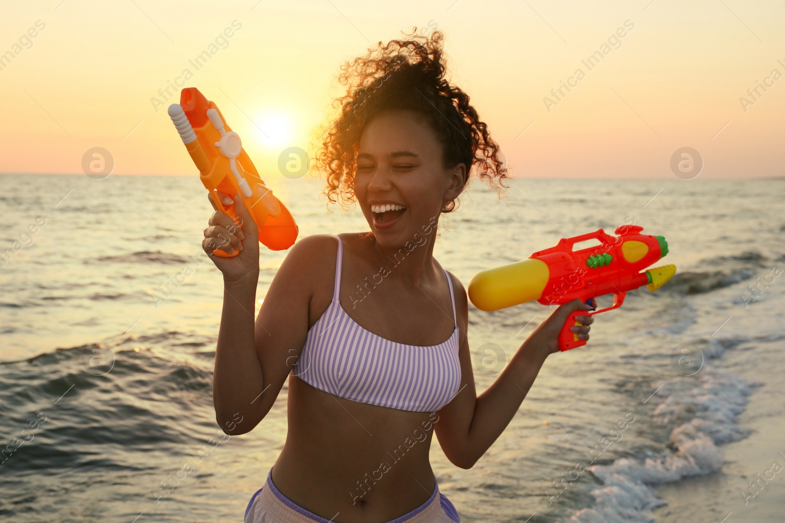 Photo of African American woman with water guns having fun on beach at sunset