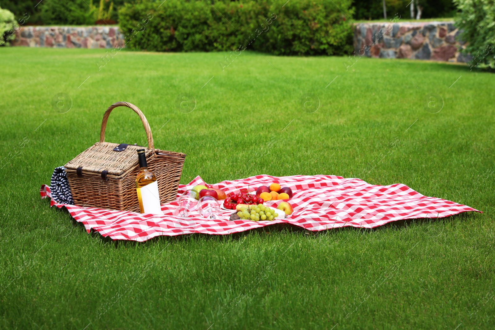 Photo of Picnic basket with products and bottle of wine on checkered blanket in garden