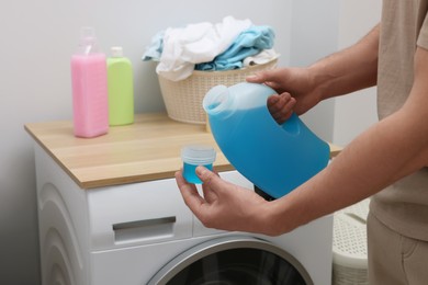 Man pouring fabric softener from bottle into cap near washing machine indoors, closeup