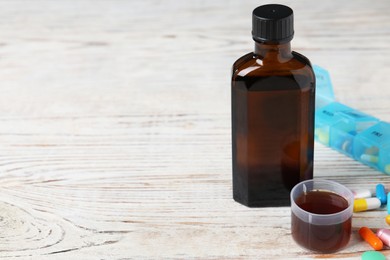 Bottle of syrup, measuring cup and pills on white wooden table background, space for text. Cold medicine