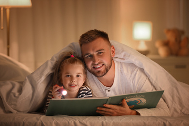 Father and daughter with flashlight reading book under blanket at home