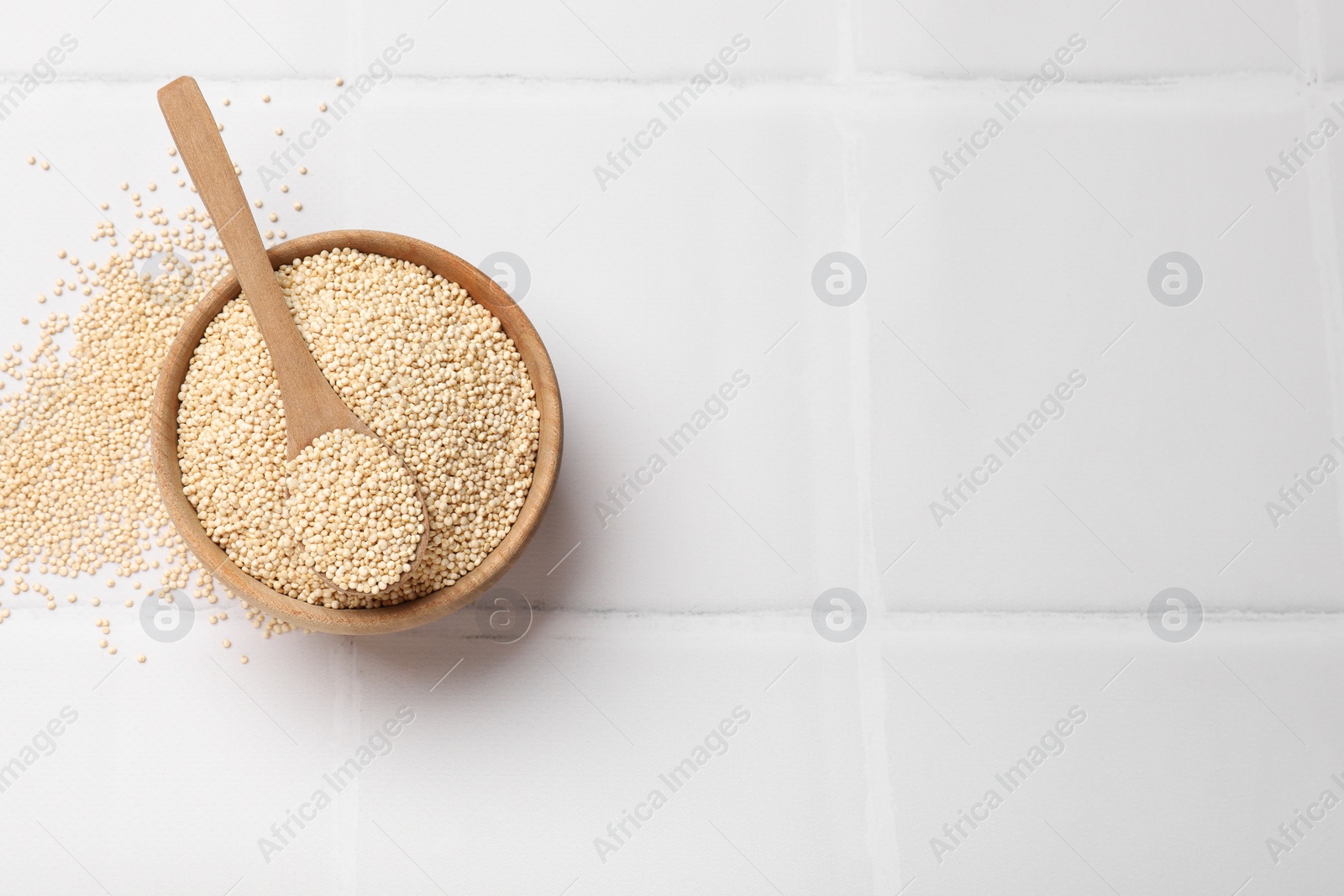 Photo of Dry quinoa seeds and spoon in bowl on white tiled table, top view. Space for text