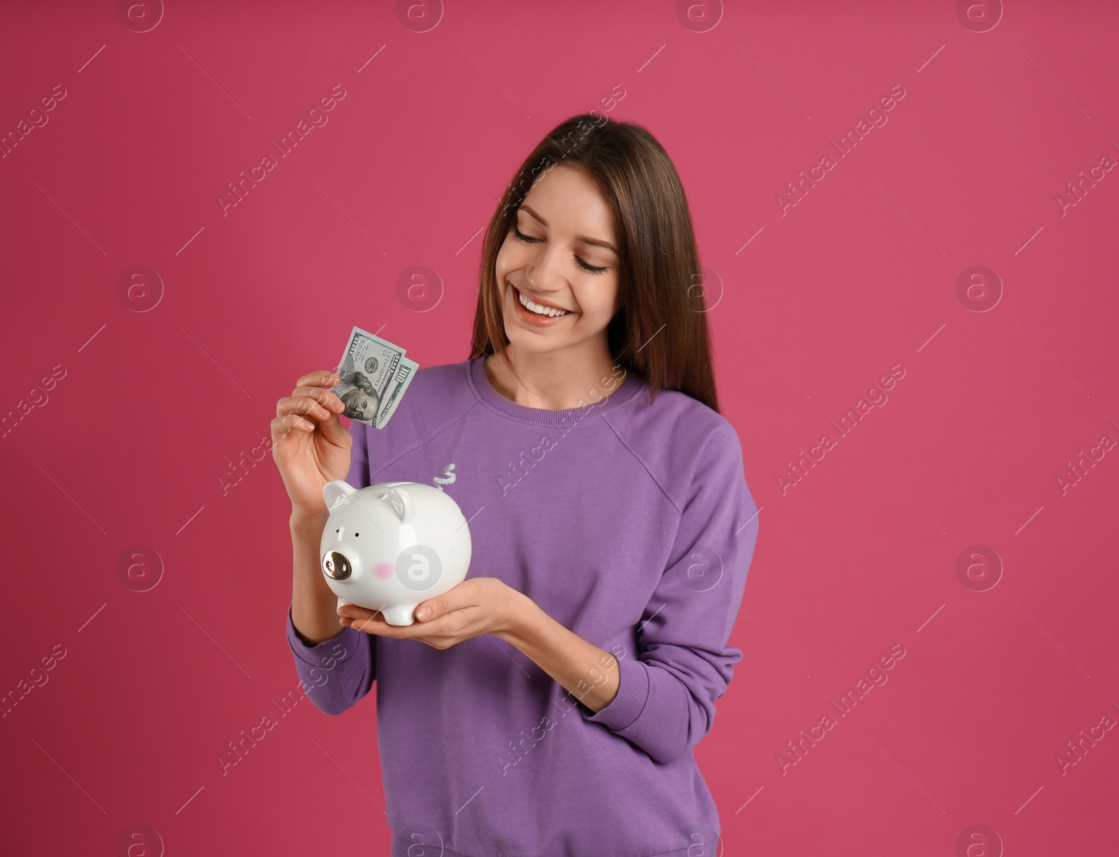 Photo of Happy young woman putting money into piggy bank on pink background, space for text