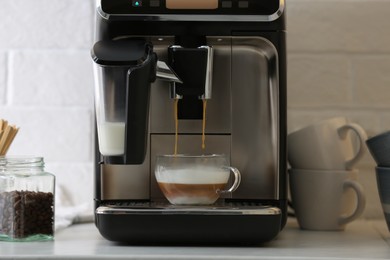 Photo of Modern espresso machine pouring coffee into glass cup with milk on white countertop in kitchen