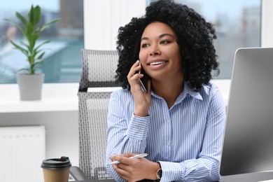 Photo of Young woman talking on phone while working with computer at table in office