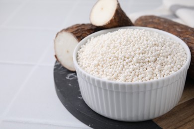 Photo of Tapioca pearls in bowl and cassava roots on white tiled table, closeup. Space for text