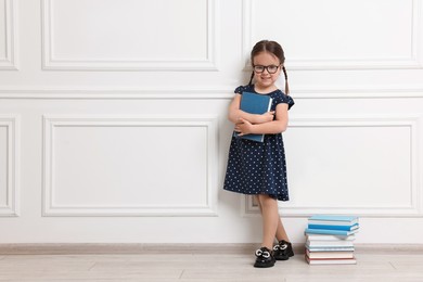 Photo of Cute little girl in glasses and stack of books near white wall. Space for text