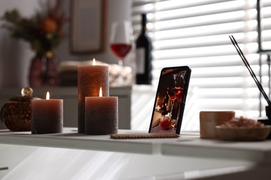 White wooden tray with smartphone, burning candles and beauty products on bathtub in bathroom