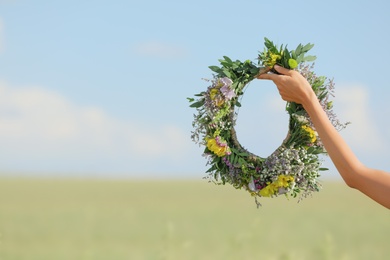 Young woman holding wreath made of beautiful flowers in field on sunny day, closeup