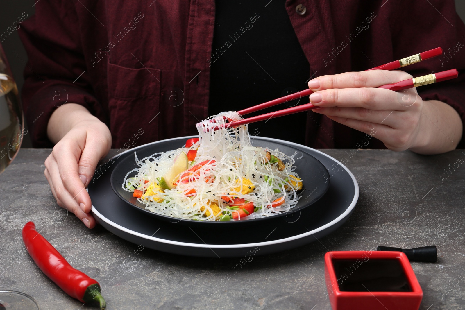 Photo of Stir-fry. Woman with chopsticks eating tasty rice noodles with meat and vegetables at grey textured table, closeup