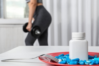 Plate of weight loss pills on table and woman with dumbbell indoors. Space for text