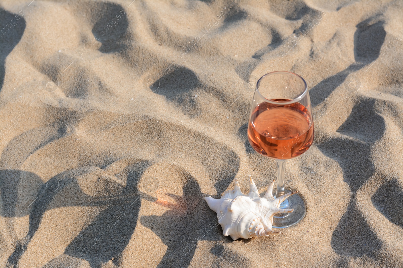 Photo of Glass of tasty rose wine and seashell on sand, space for text