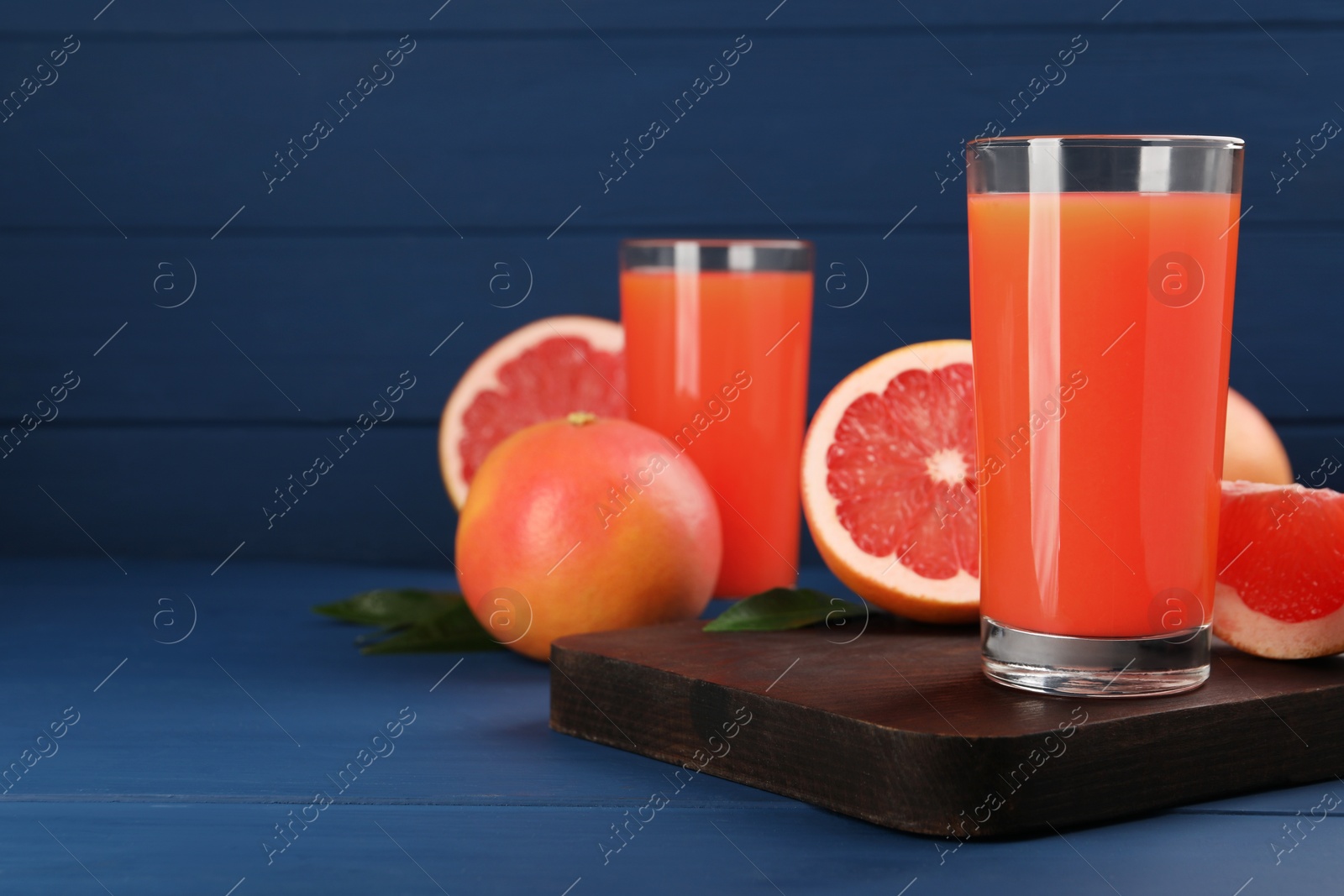 Photo of Tasty grapefruit juice in glass and fresh fruits on blue wooden table. Space for text