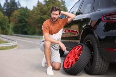 Photo of Tire puncture. Stressed man with new wheel near car on roadside outdoors