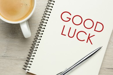 Image of Notebook with phrase GOOD LUCK, pen and cup of coffee on white wooden table, flat lay