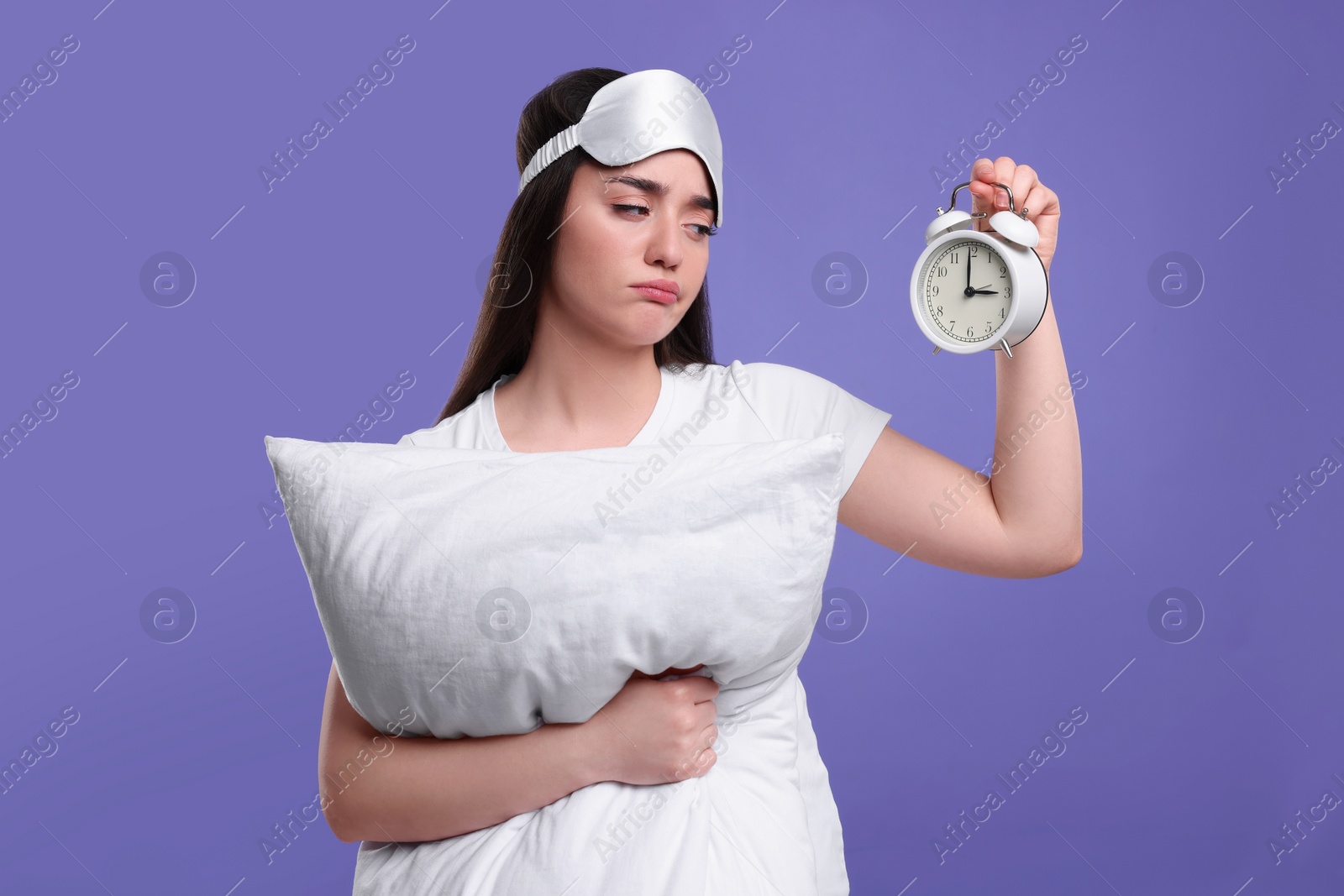 Photo of Tired young woman with sleep mask, alarm clock and pillow on purple background. Insomnia problem