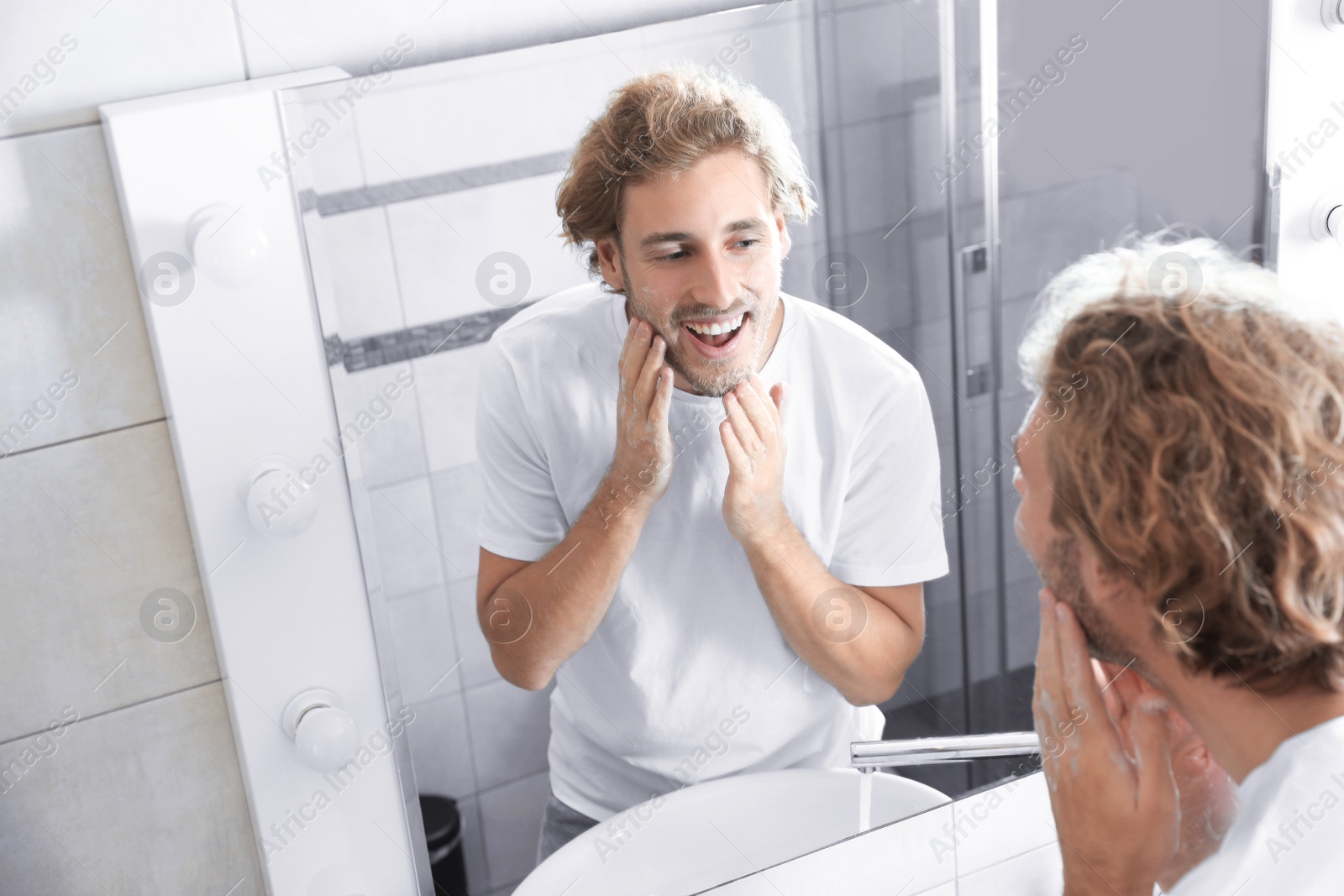 Photo of Young man washing face with soap near mirror in bathroom