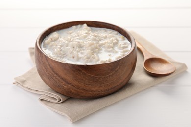 Tasty boiled oatmeal in bowl and spoon on white wooden table, closeup