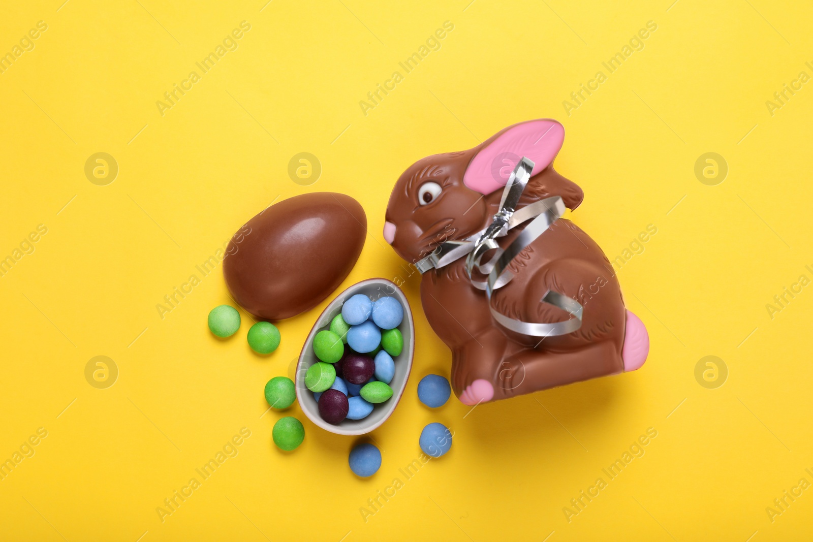 Photo of Chocolate Easter bunny, halves of egg and candies on yellow background, flat lay