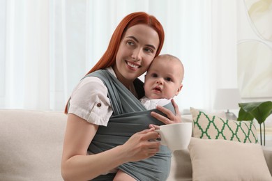Mother with cup of drink holding her child in sling (baby carrier) at home