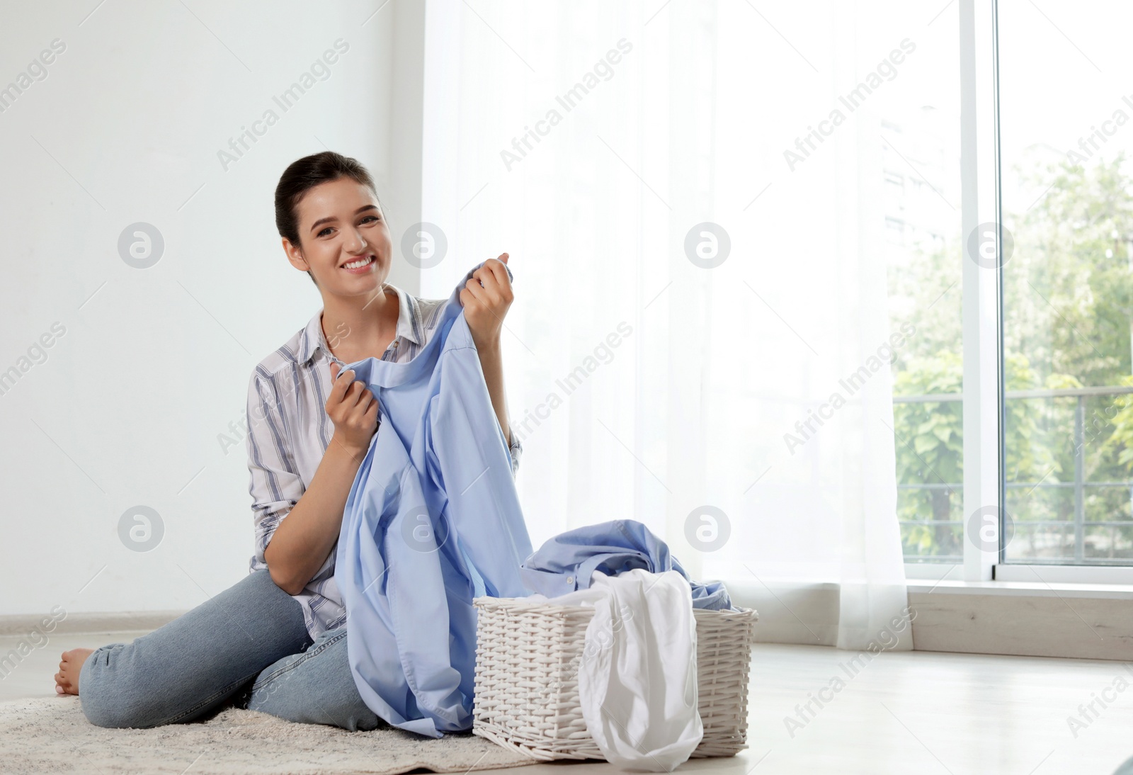 Photo of Young woman with laundry basket and clean clothes sitting on floor indoors. Space for text