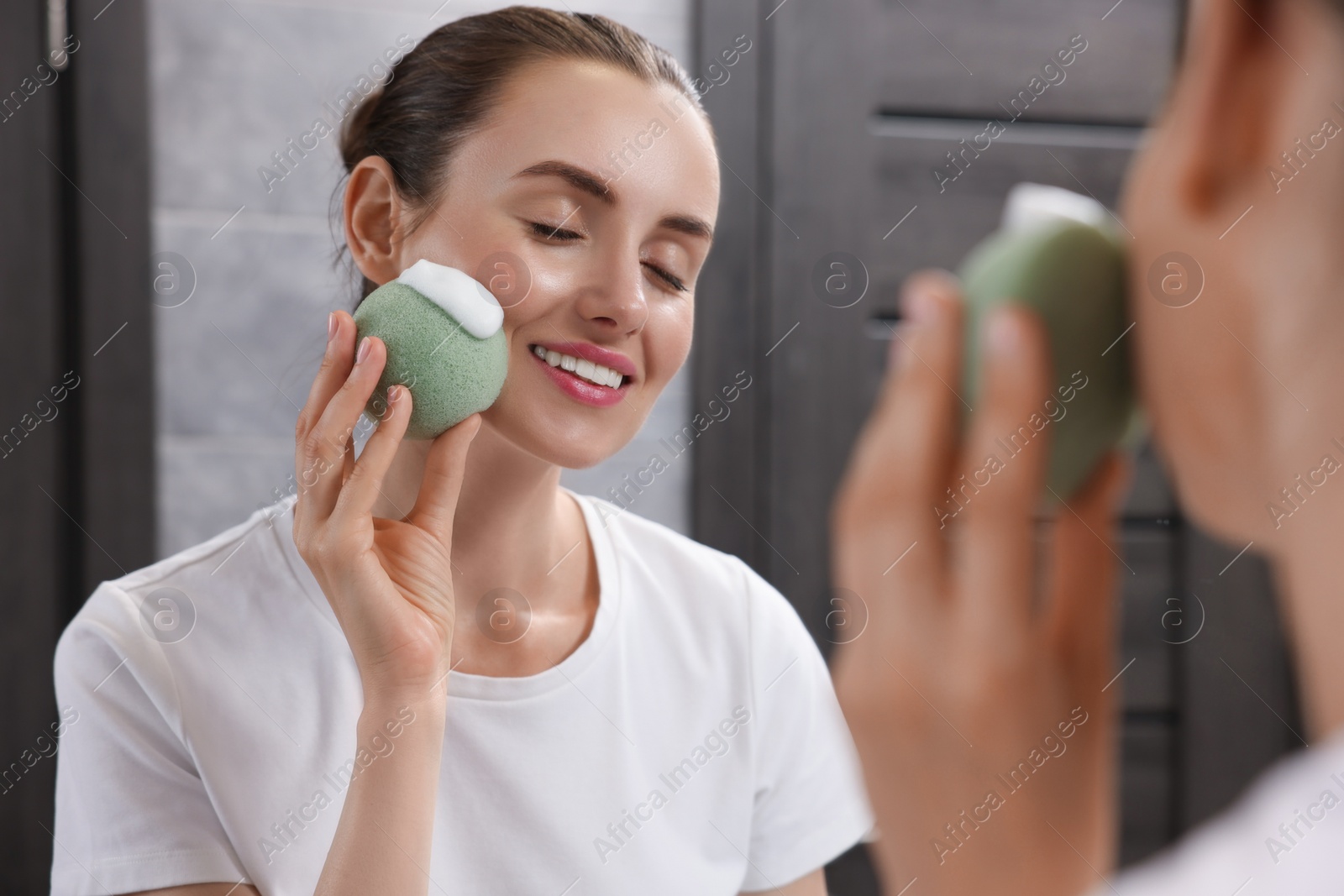 Photo of Happy young woman washing her face with sponge near mirror in bathroom