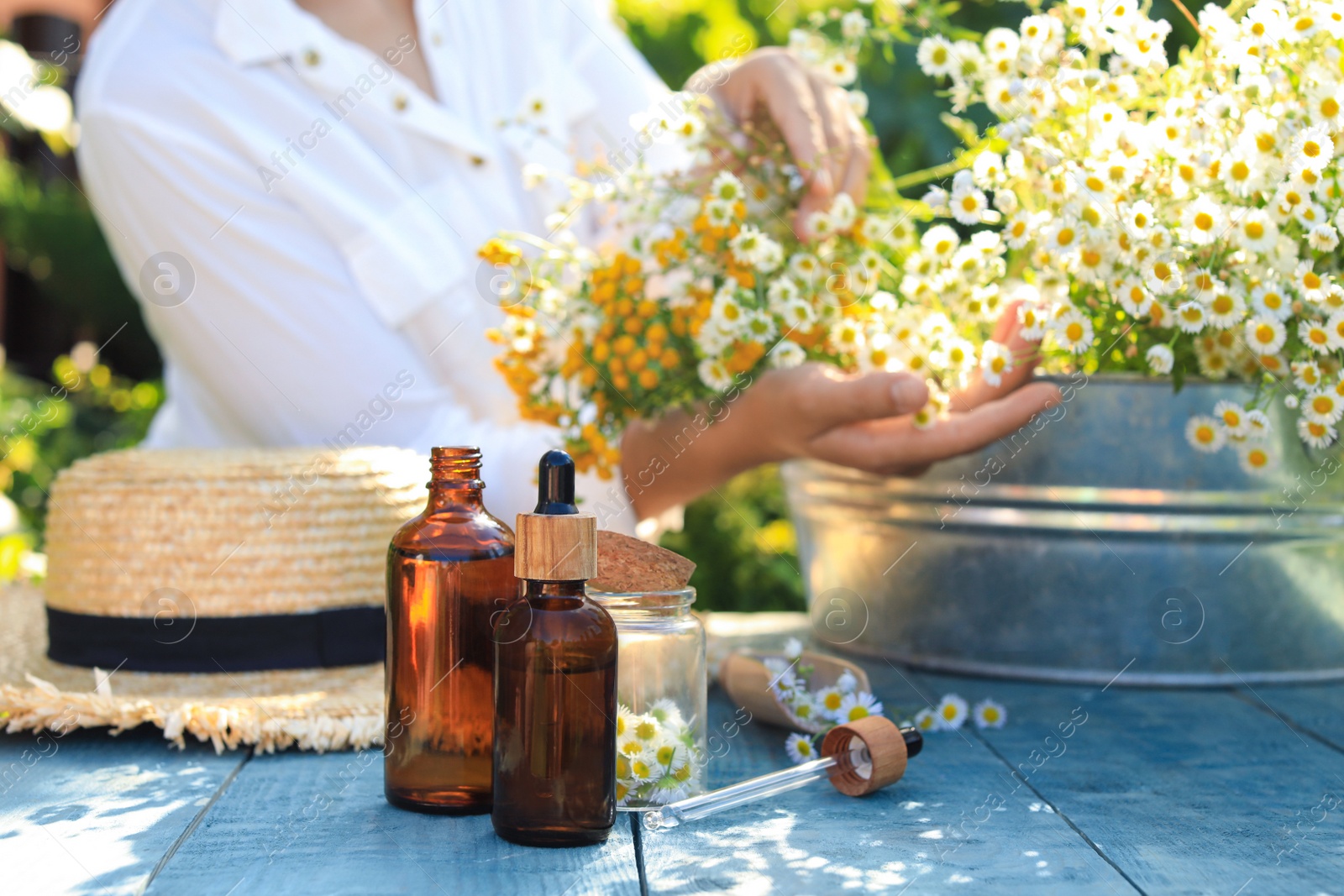 Photo of Woman with flowers near table outdoors, focus on bottles of chamomile essential oil