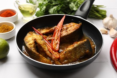 Photo of Tasty fish curry in frying pan and ingredients on white tiled table, closeup. Indian cuisine